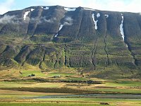 A farm stands on the narrow plain below erosion patterns on the green mountainside.