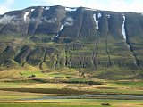 A farm stands on the narrow plain below erosion patterns on the green mountainside.
